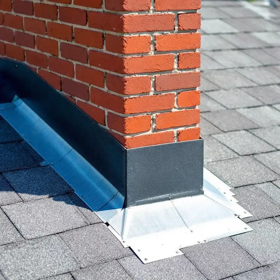 Red brick chimney passing through a shingle roof, metal flashing worked into the brickwork and lapped over the shingles at its base, a small raised sloped cricket built behind it on the uphill side