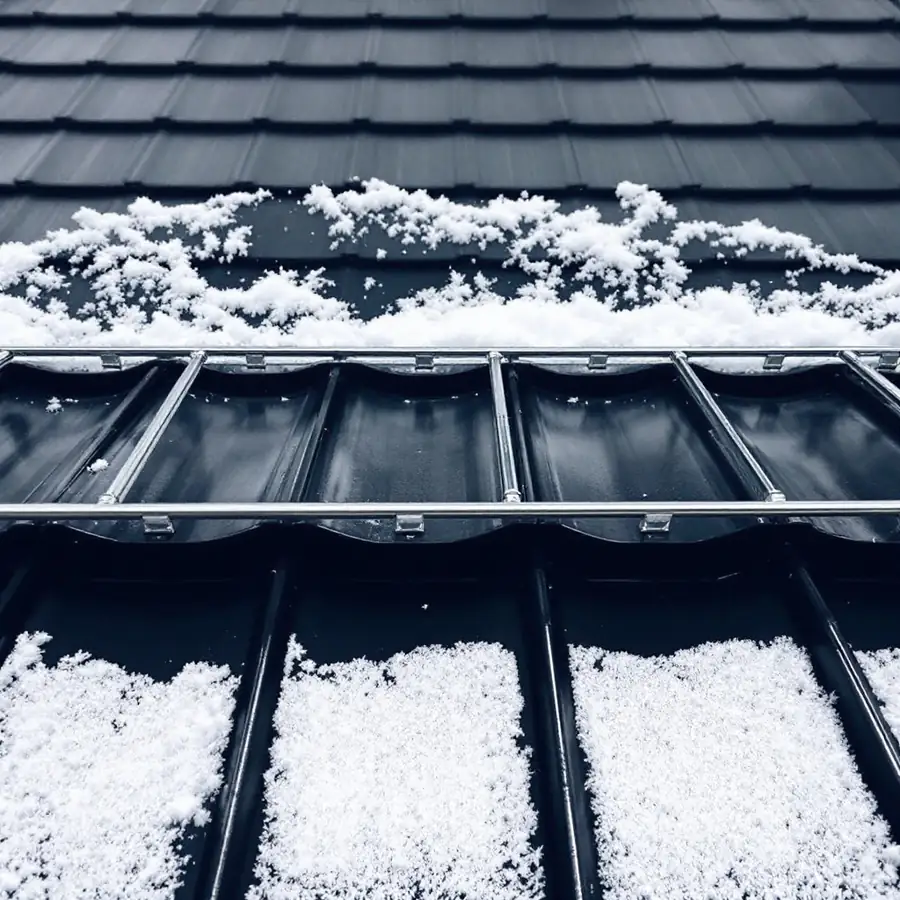 Dark metal roof with a horizontal row of metal snow retention bars mounted across the panels, snow banked behind the bars and clear panel below them
