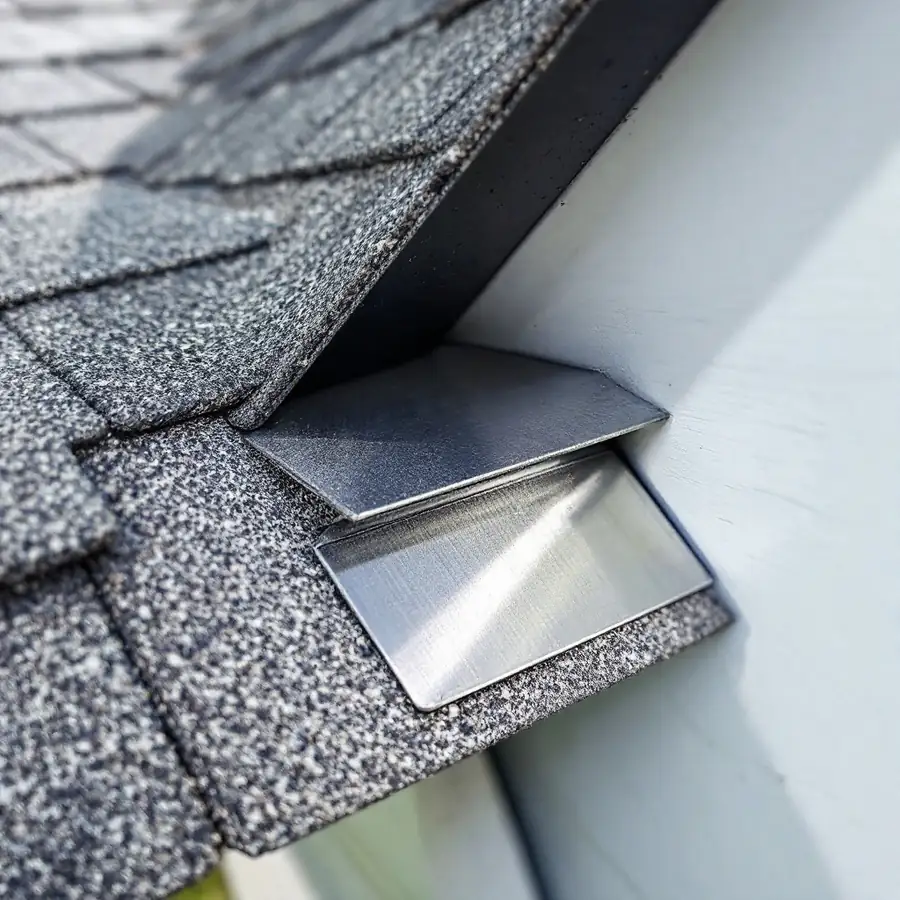 Junction where a shingle roof slope meets a vertical wall, showing overlapping rectangular metal step flashing pieces tucked between the shingle courses