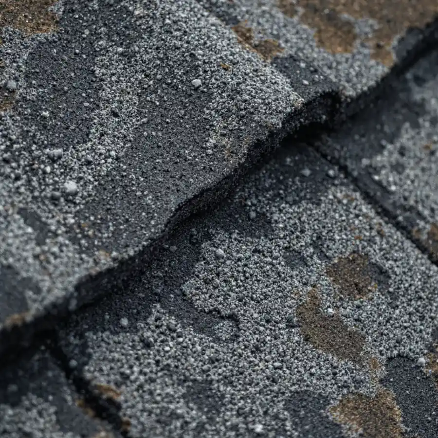 Extreme close-up of weathered asphalt shingles where mineral granules have worn away in patches, exposing smooth black bitumen beneath the remaining grey granules