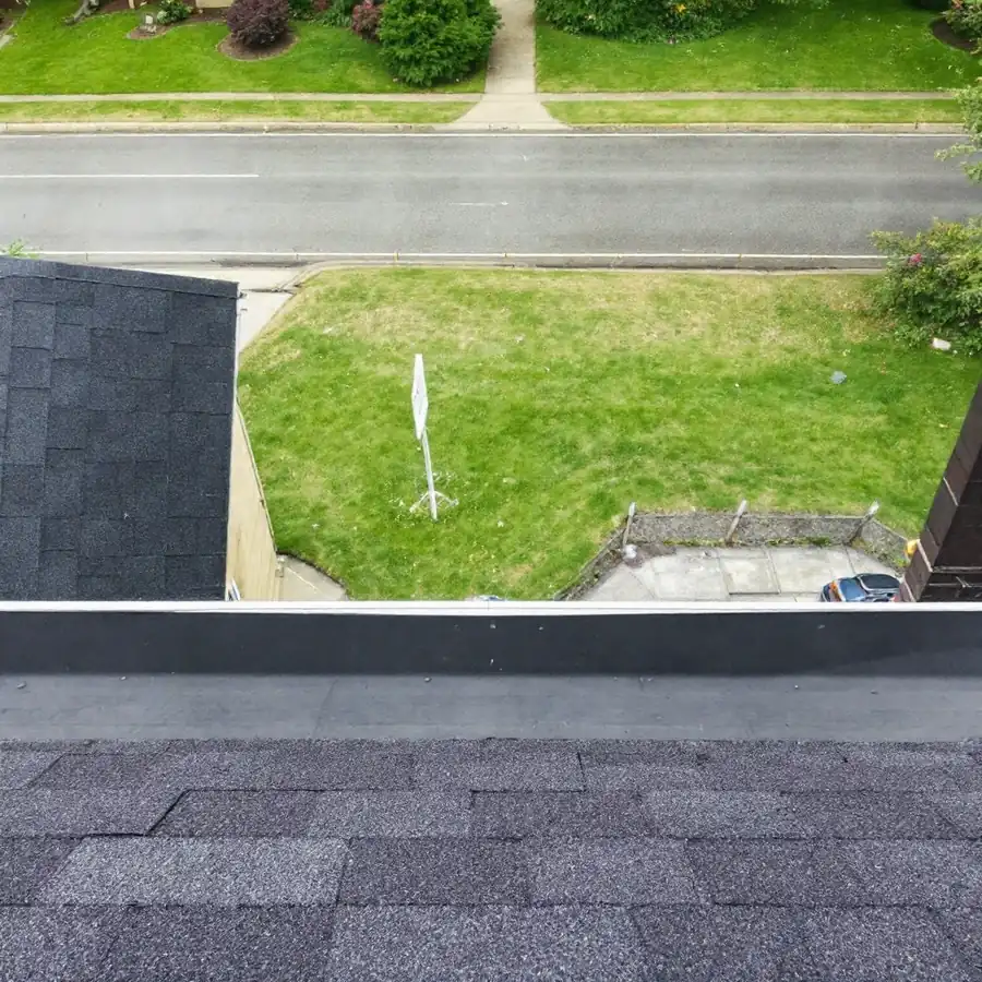 View from the edge of a roof looking straight down past the gutter and drip edge to the lawn and path below, with dark shingles in the foreground