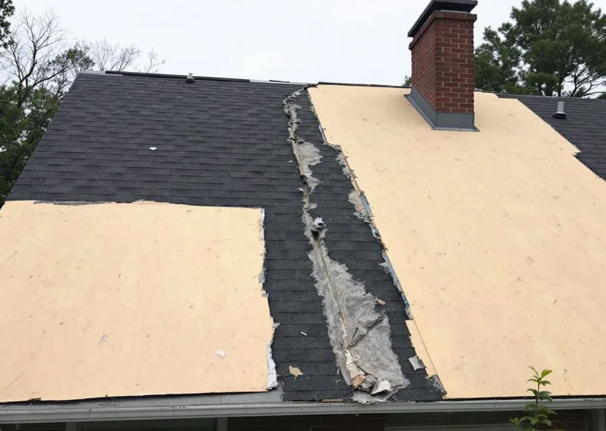 A house roof in the middle of a tear-off: the left half stripped to pale plywood decking, the right half still carrying dark asphalt shingles, with torn felt along the ragged line between them and a brick chimney above