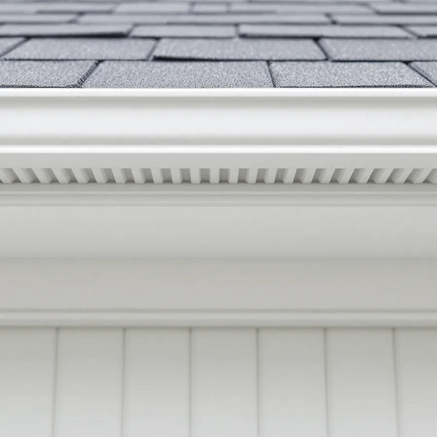 Looking up at the underside of a roof eave showing a continuous white vented soffit panel with fine slots, white fascia board and the edge of grey shingles above