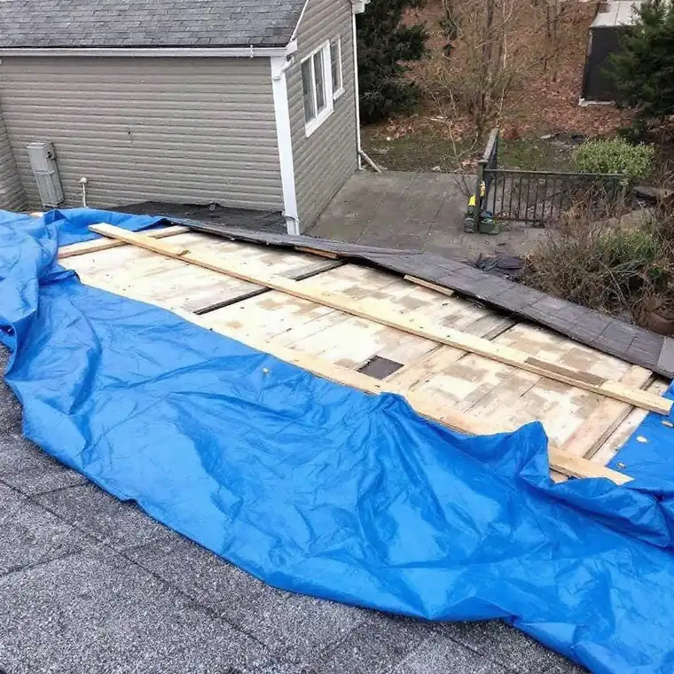 Blue emergency tarp stretched across a damaged roof slope and held down with wooden battens, exposed decking visible alongside